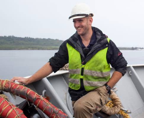 A smiling man wearing a white hard hat, a reflective yellow vest, and tan trousers stands on a boat, holding a thick rope with water and greenery in the background—capturing the spirit of teamwork found in our About Us story.